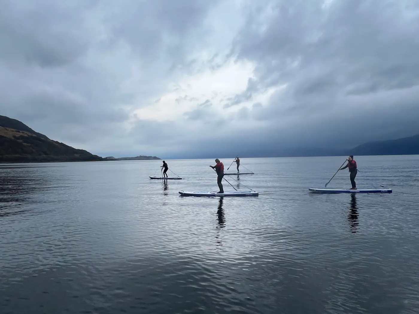 Four paddleboarders crossing a calm Scottish loch beneath a stormy sky on a Sup2summit expedition