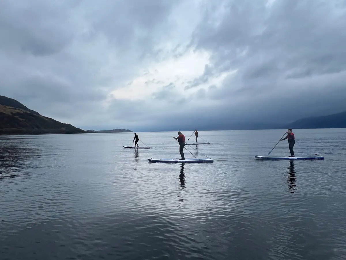Four paddleboarders crossing a calm Scottish loch beneath a stormy sky on a Sup2summit expedition