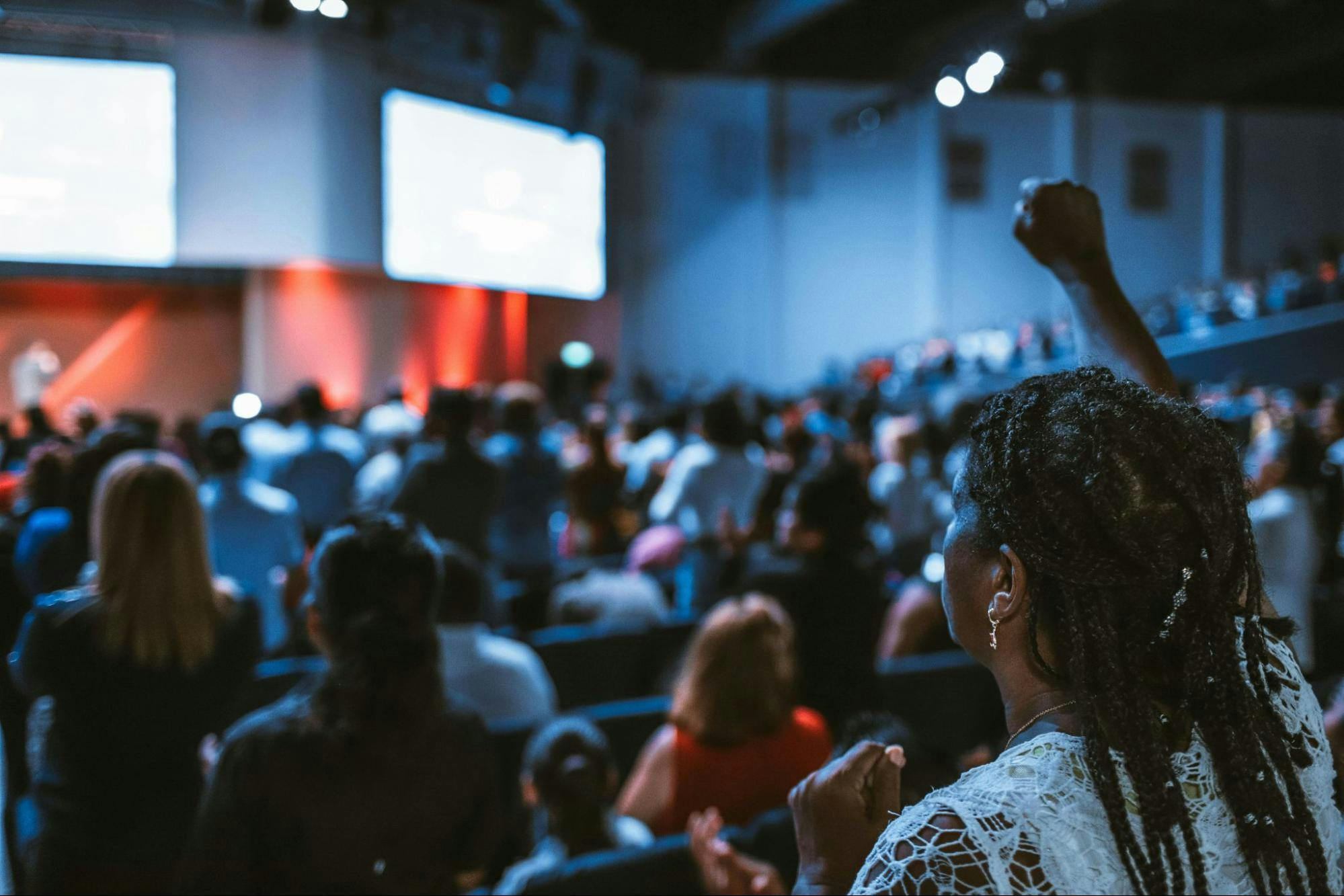 A crowd in an auditorium watches a presentation, with one person in the foreground raising their fist.