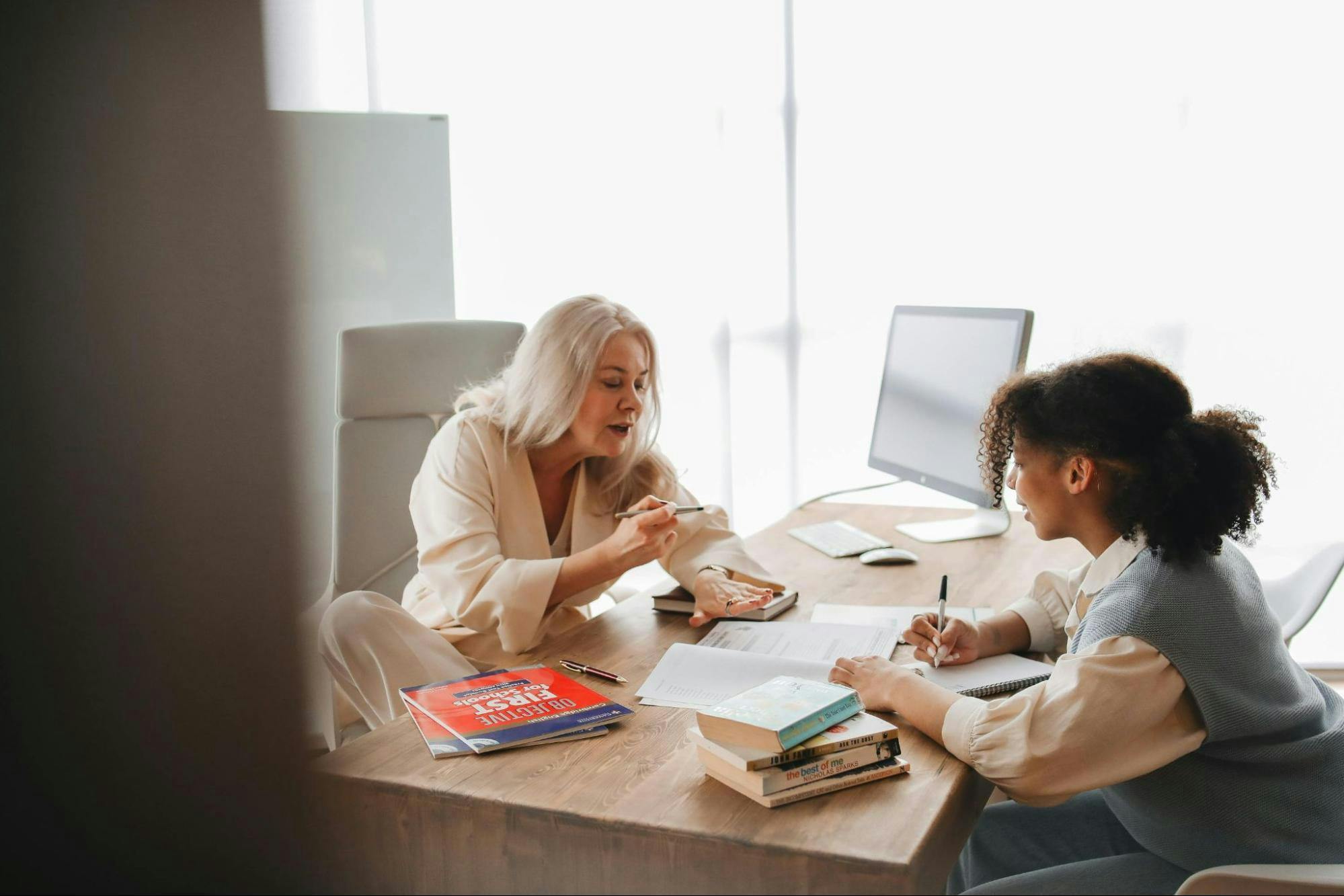 Two women sit at a desk with books and documents, one with gray hair speaking, and the other with curly hair taking notes. A computer and an empty chair are visible in the background.