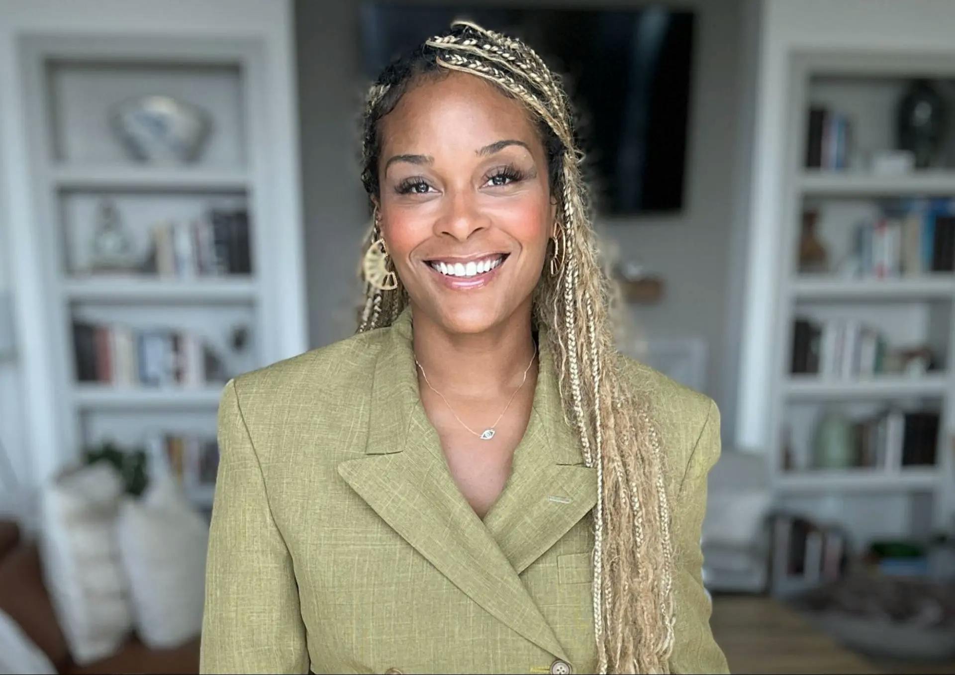 Erin Patten with long blonde braids, wearing gold earrings and a light green blazer, smiles at the camera. The background features bookshelves and a cozy, well-lit living room.