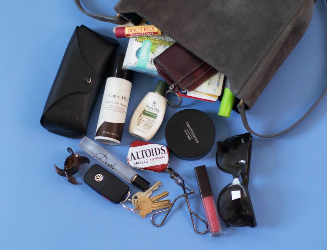 Image of esalon employee's dark handbag tipped over with esalons camo mist, sunglasses, lip gloss, eyelash curler, aveeno hand lotion, sunglasses case, burt's bees lip balm, dolce and gabbana perfume roller, and a hair clip 