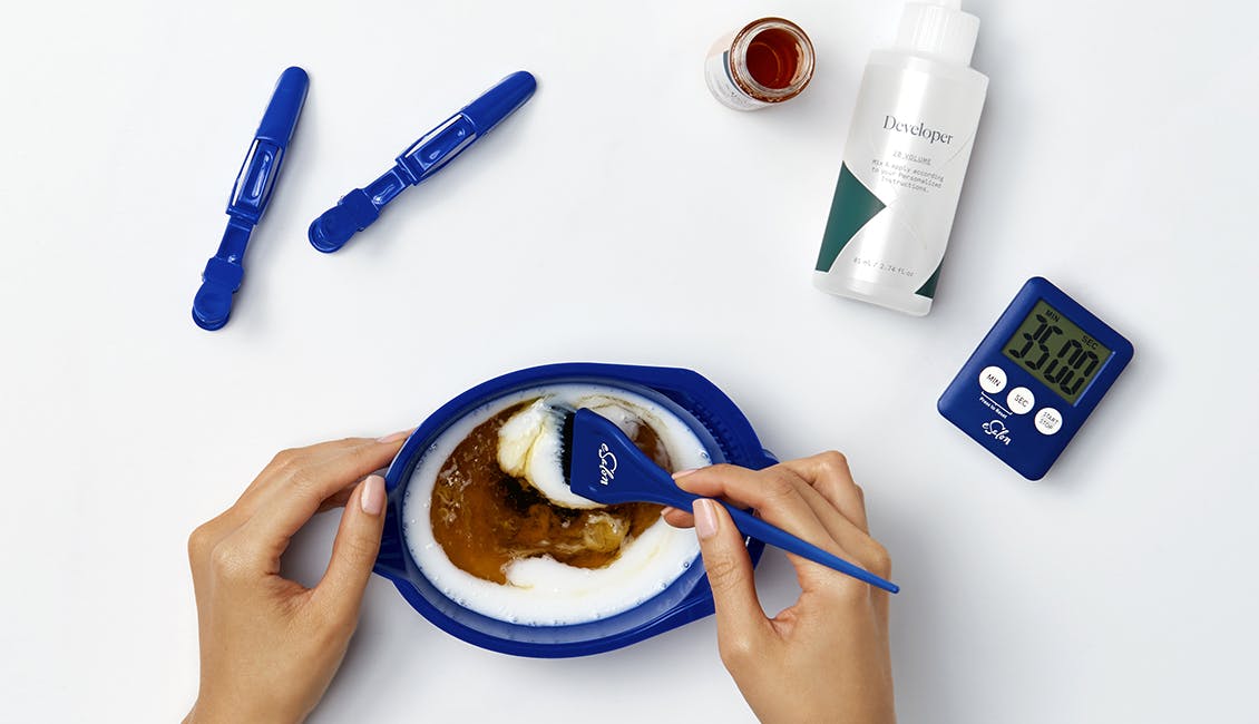 Image of hands mixing custom hair color with developer in a mixing bowl with two hair clips, a timer, and color bottle set out on the table