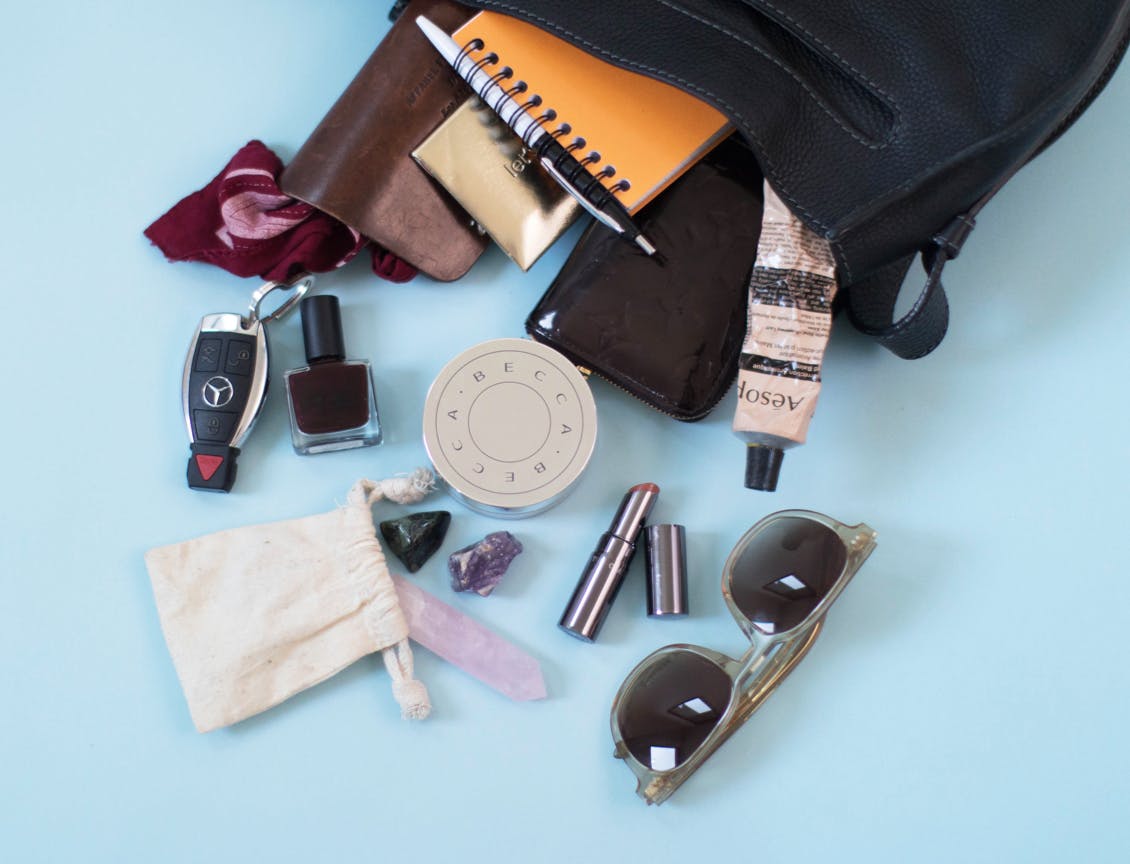 Image of eSalon employee courtney's black handbag with her favorite beauty essentials and misc items including lipstick, sunglasses, car keys, notebook and pencil, loose face powder, bag of spilled crystals, and aesop hand lotion tube 