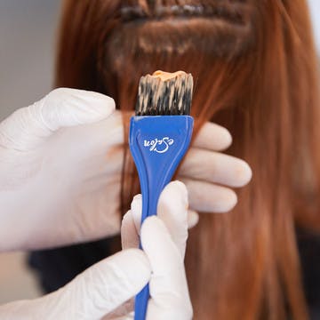 Image of someone with gloved hands and a an eSalon tint brush with hair color on it applying color to someone else's hair
