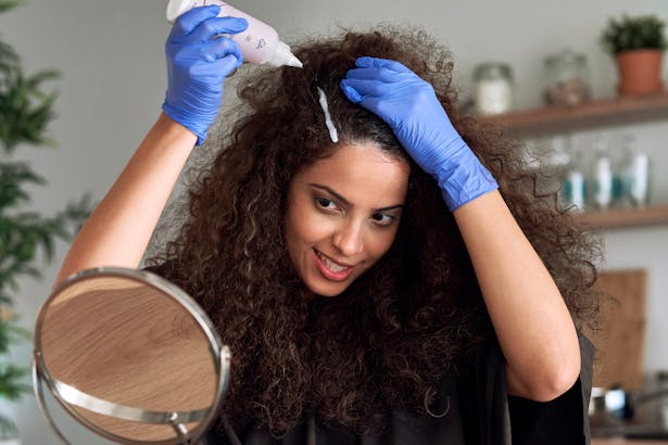 Woman with brown curly hair coloring her hair in the kitchen. 