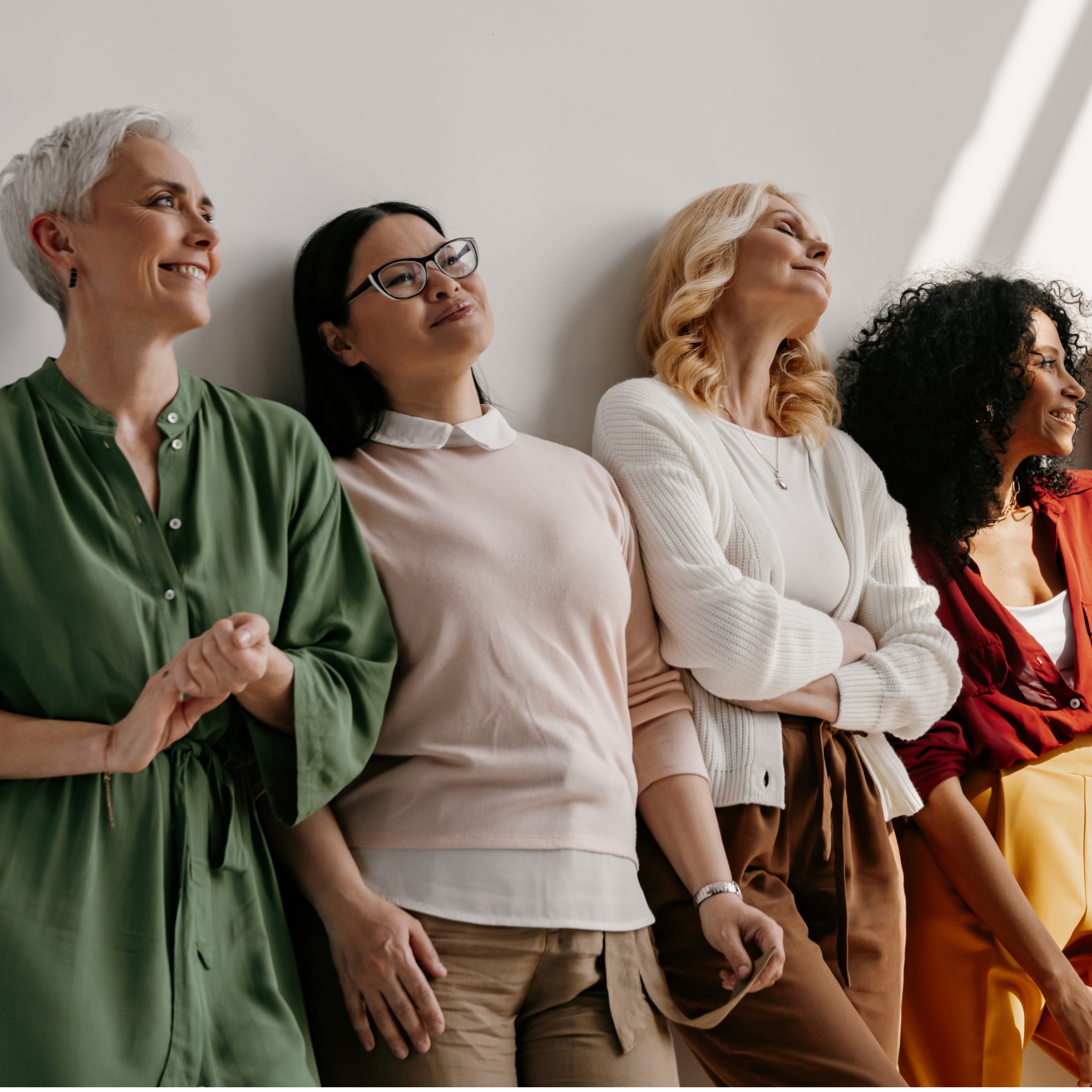 Group of women of varying ages and hair types leaning against a sunlit wall.