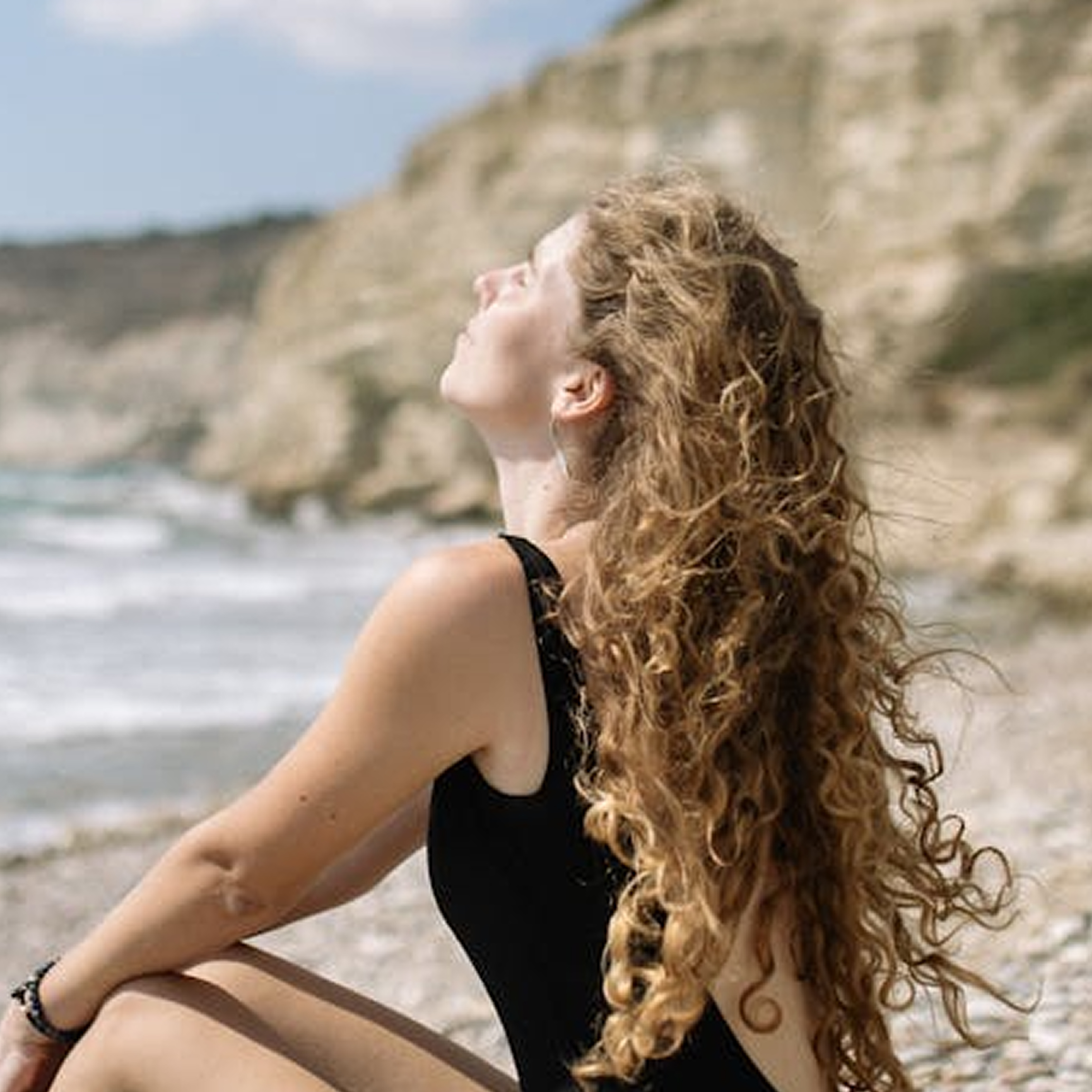 Woman sitting on the beach with long, flowy, curly blonde hair. 