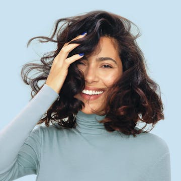 Image of woman looking thrilled with esalon custom hair color in brunette on a blue background and wearing a blue turtleneck