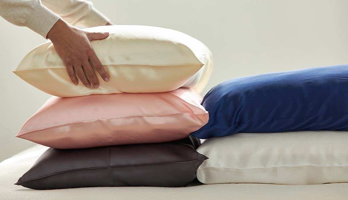 Image of five stacked pillows with eSalon x Avocado Silk Pillowcases on in five different colorways: ivory, pink, charcoal, navy, and white. Three are stacked on top of each other, with the remaining two stacked on the side. Hands are stacking the pillows. 