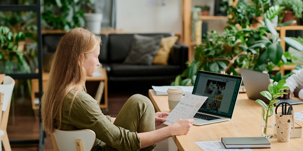 Woman sitting at a desk, reading a paper in front of a laptop. Background with lots of plants and a couch.