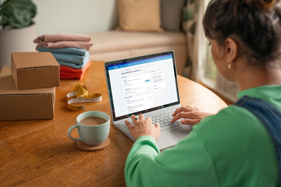 woman sat at table using laptop with drink and parcels next to her