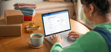 woman sat at table using laptop with drink and parcels next to her