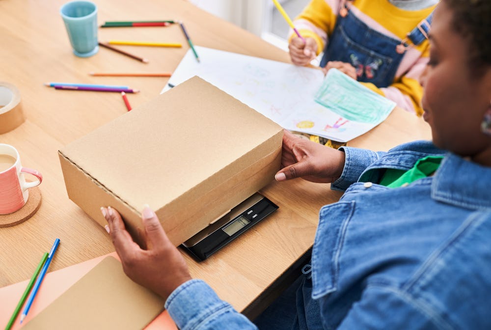 woman placing cardboard box on weighing skills while child draws next to her
