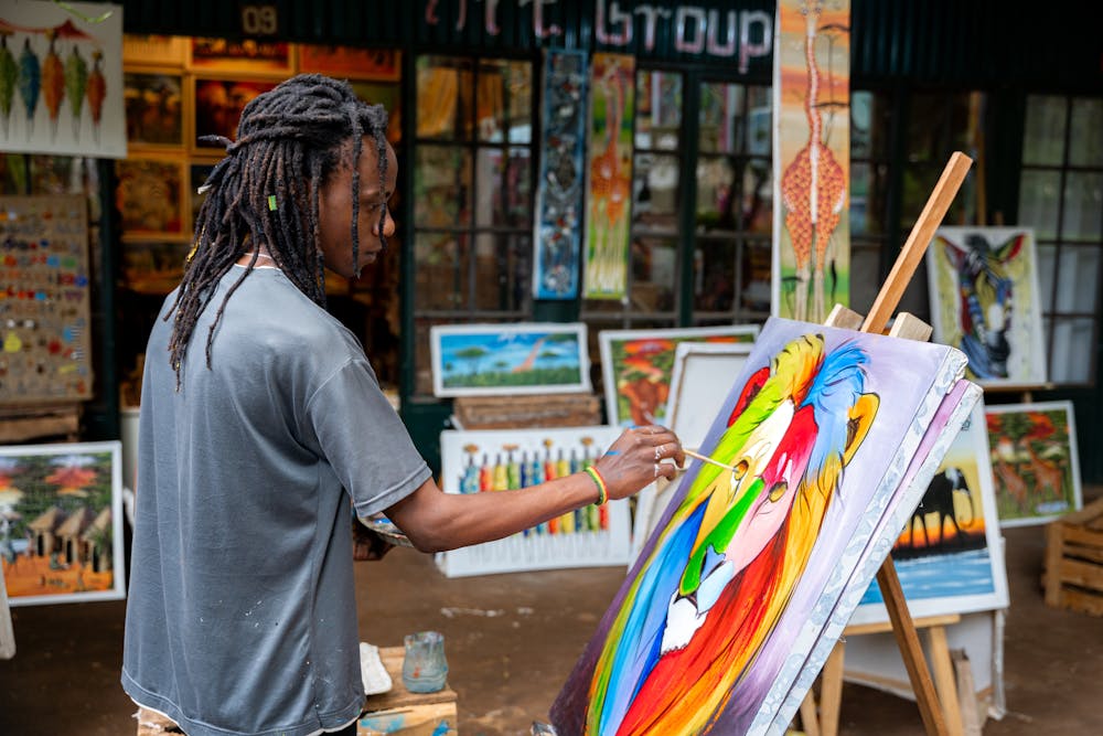 An artist is painting a multi-coloured lion on canvas with an easel propping it upright in a street. You can see colourful paintings displayed in the shop window behind