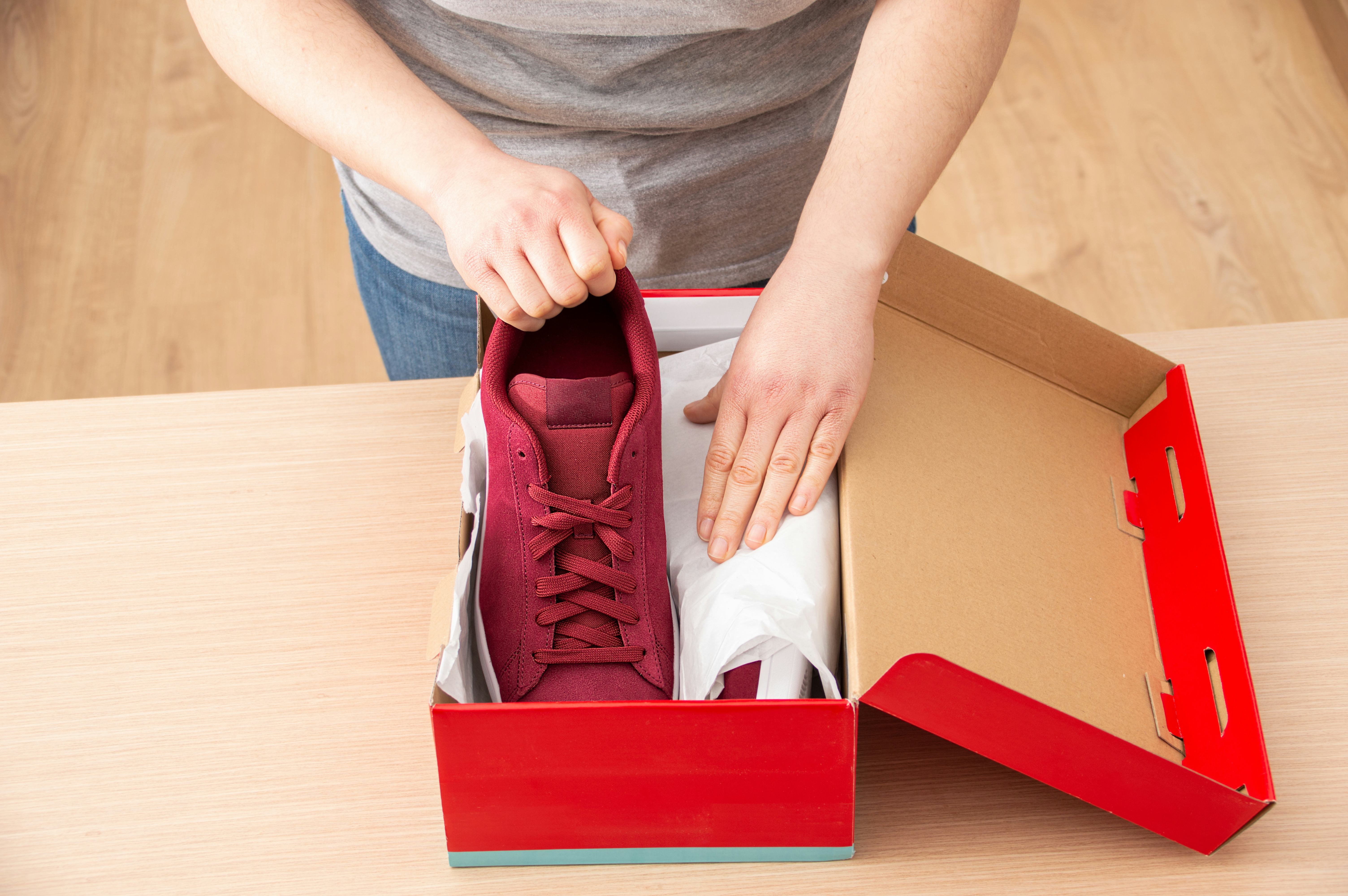 A close-up of a customer standing and facing the camera, focusing on a red shoe box on top of a table. They are placing a pair of red trainers inside the packaging.