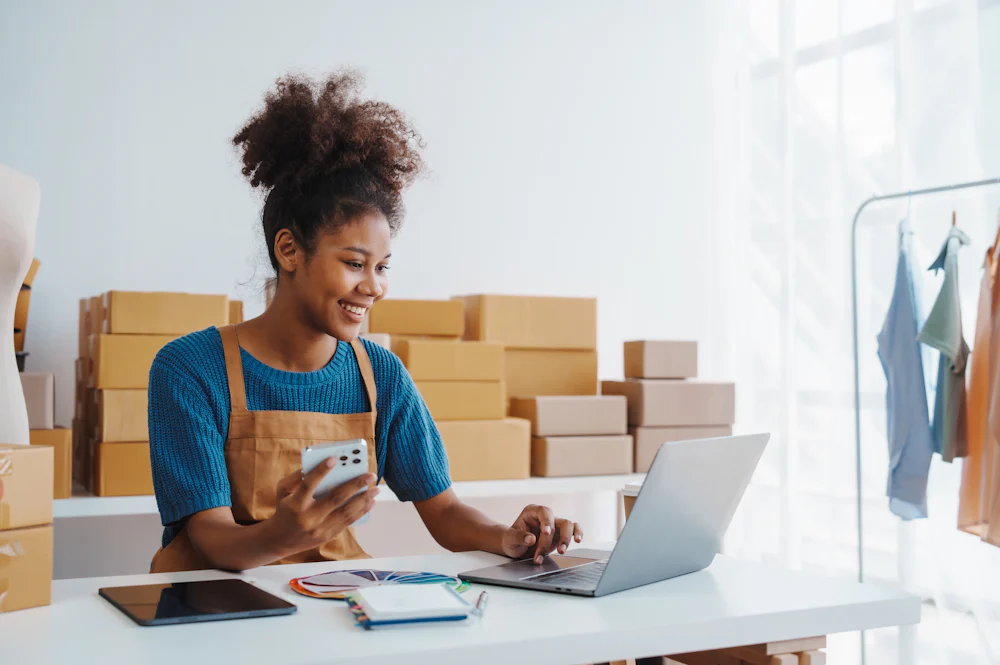 A side profile view of a business owner sitting at a desk in their office using their laptop and smiling. Stacked parcel boxes can be seen in the background.