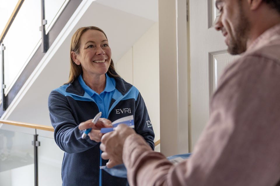 An Evri Courier handing a customer a blue slip to confirm the details of a doorstep parcel collection. The transaction is taking place in a block of flats