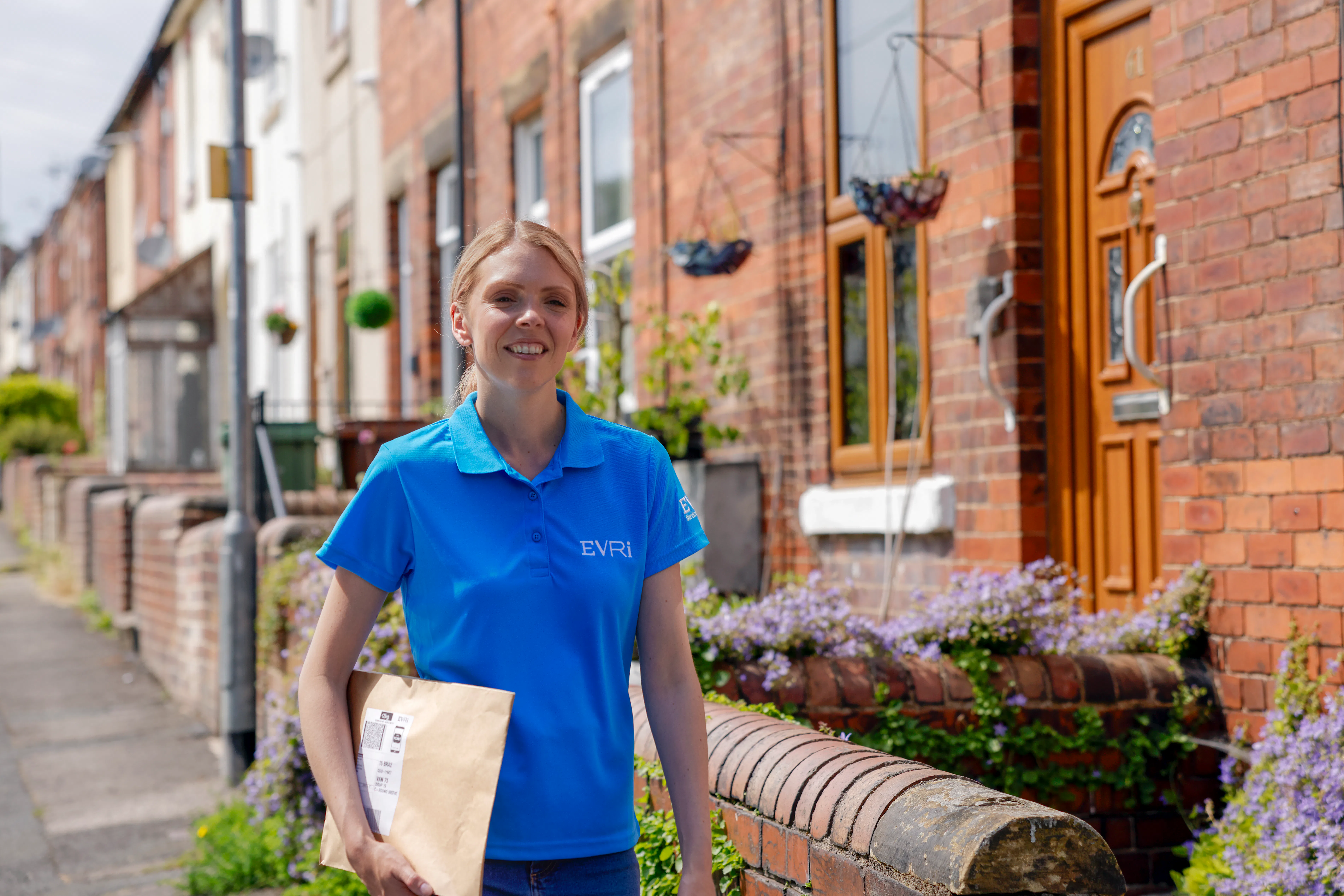 An Evri courier is walking down a street of terraced houses carrying a parcel. The greenery and architecture suggests a rural British town or village.