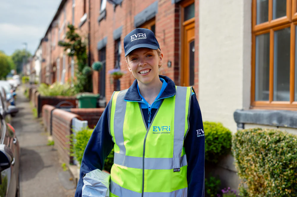 An Evri courier in high vis is smiling posing towards the camera with a parcel under their arm. The backdrop is a street of terraced houses in a rural British neighbourhood.