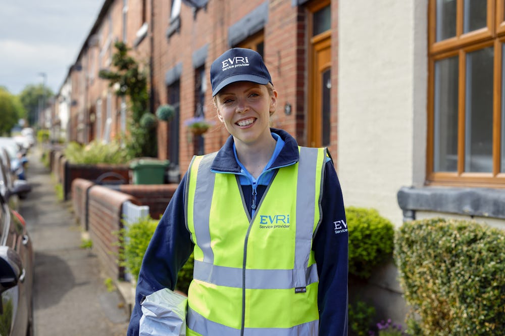 An Evri courier in high vis is smiling posing towards the camera with a parcel under their arm. The backdrop is a street of terraced houses in a rural British neighbourhood.
