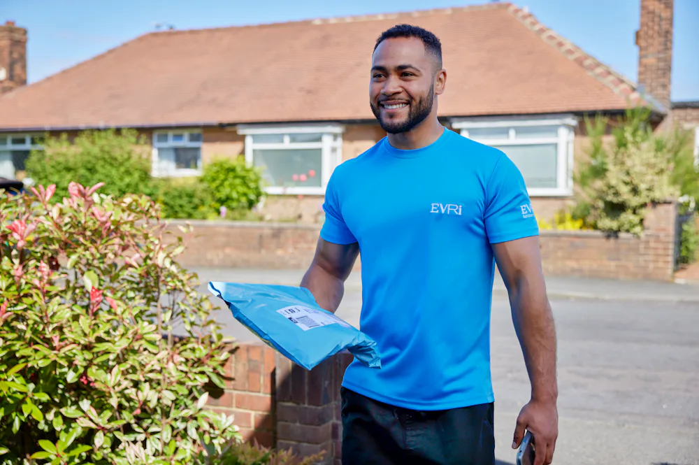 An Evri Courier is approaching the doorstep of a house in a residential area. He is carrying a single parcel, ready to deliver it.