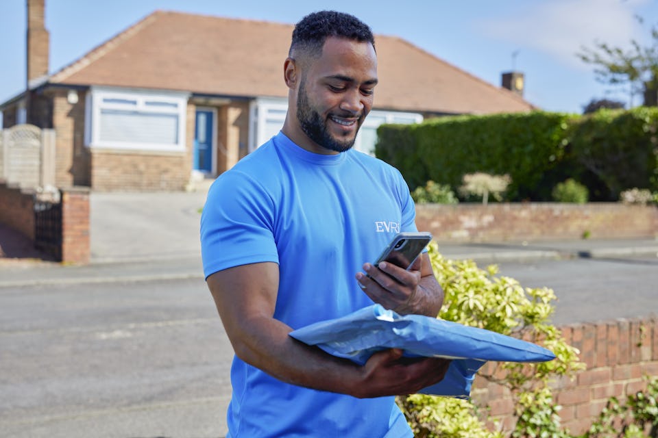 An Evri Courier is scanning the barcode of a single parcel as he approaches the doorstep of a home in a residential area. He is there to make a delivery.