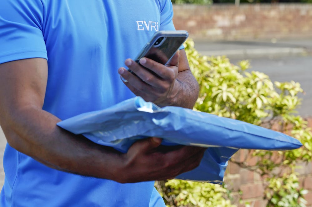 An Evri Courier is scanning the barcode of a single parcel as he approaches the doorstep of a home in a residential area. He is there to make a delivery.