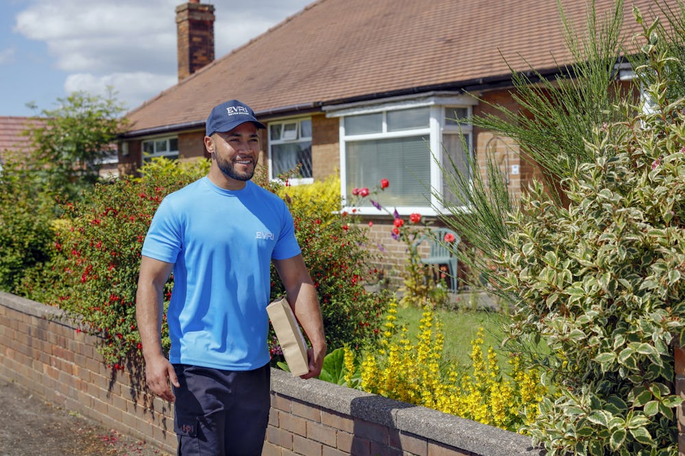 An Evri Courier is walking down the street in a residential area with a parcel under his arm.