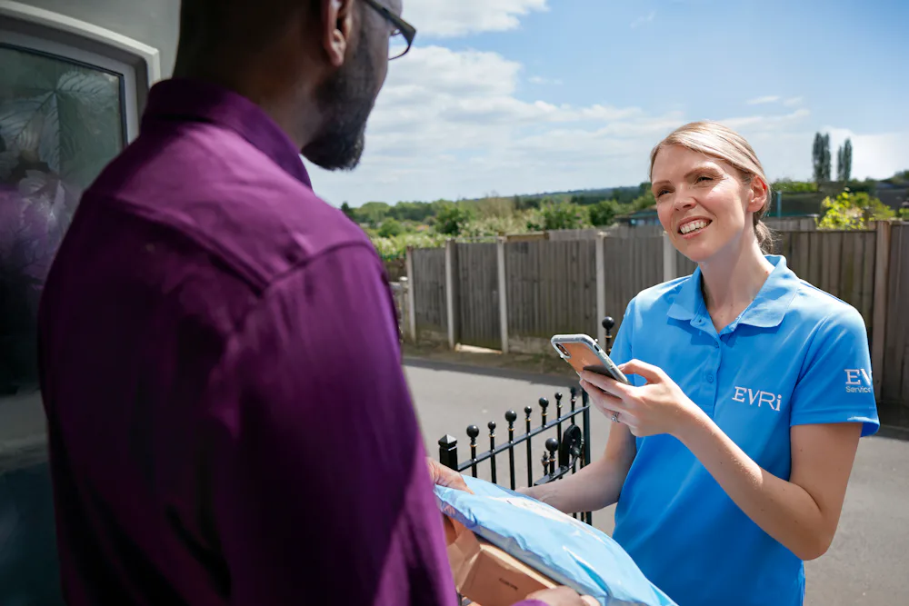 A female courier is taking a photo confirmation of a parcel delivery whilst handing the parcel to the recipient. They are exchanging in friendly dialogue whilst the photo is being taken.