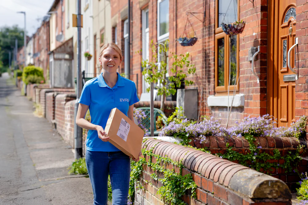 An Evri courier is walking down a street of terraced houses carrying a parcel. The greenery and architecture suggests a rural British town or village.