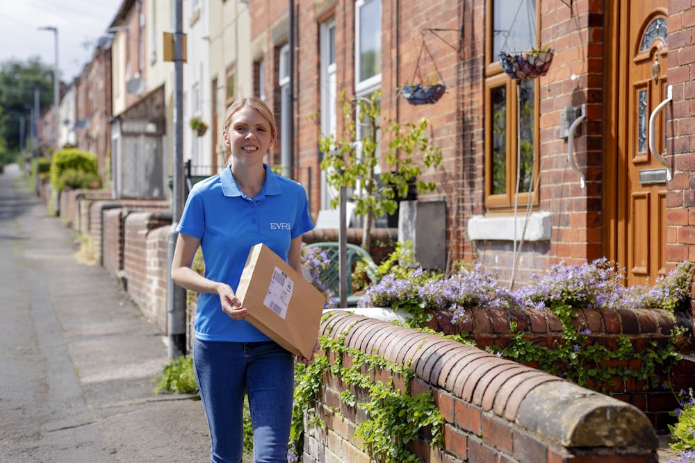 An Evri courier is walking down a street of terraced houses carrying a parcel. The greenery and architecture suggests a rural British town or village.