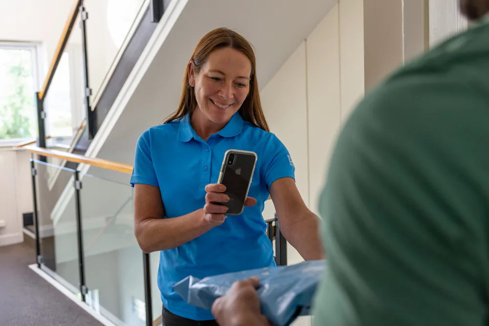 An Evri Courier is handing a parcel to a recipient at their doorstep in a block of flats. The courier is taking photo confirmation to evidence the transaction.