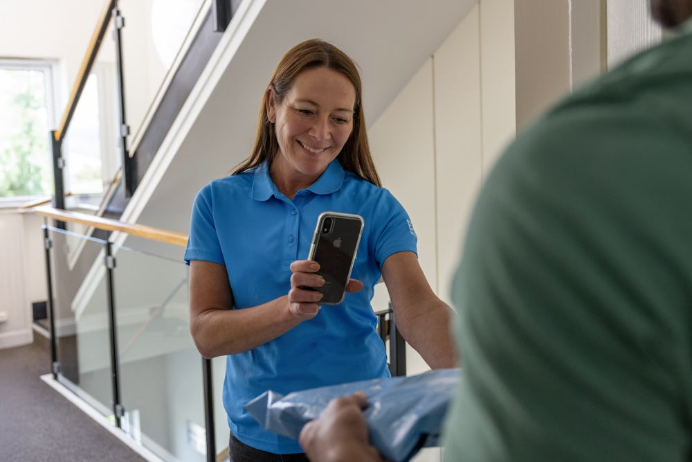 An Evri Courier is handing a parcel to a recipient at their doorstep in a block of flats. The courier is taking photo confirmation to evidence the transaction.
