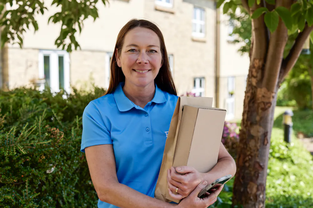 An Evri courier smiling and posing for the camera with two parcels in their grasp. The backdrop is a suburban British neighbourhood with plenty of greenery.
