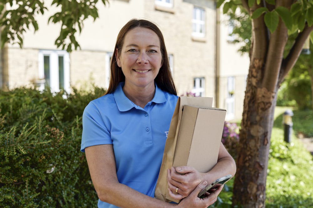 An Evri courier smiling and posing for the camera with two parcels in their grasp. The backdrop is a suburban British neighbourhood with plenty of greenery.