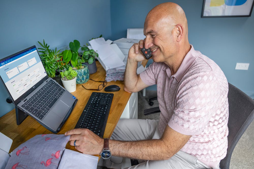Business owner sat at a desk in their home office taking a phone call and using their laptop to book multiple parcels