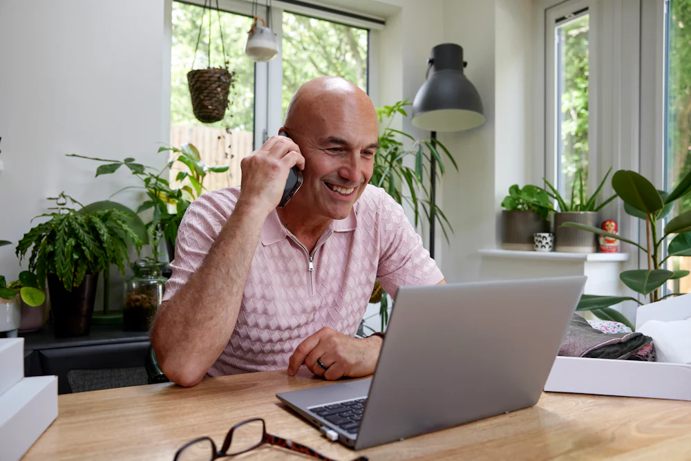 Customer sat at a desk in their office taking a phone call and looking at a laptop.