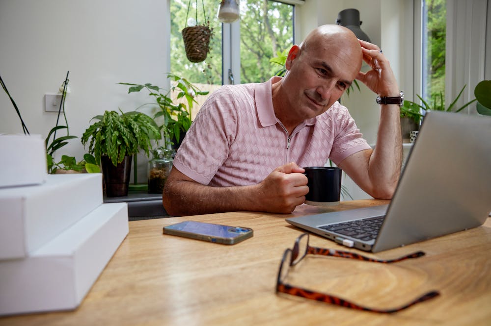 Customer sat at a desk in their office using a laptop with multiple parcels packaged next to them. One hand is positioned against their head to show concentration, the other is holding a mug.