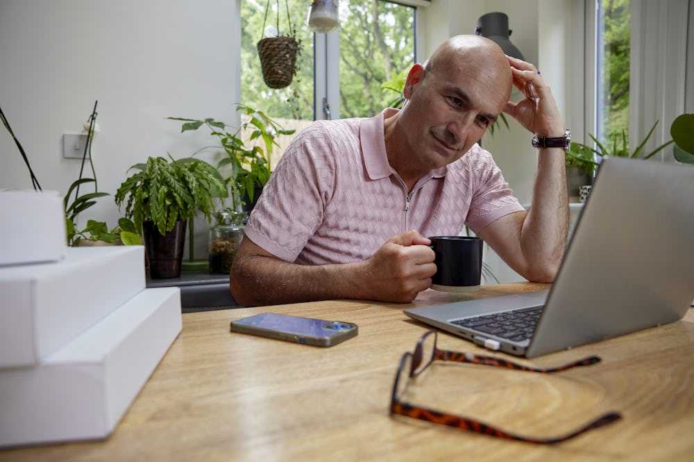 Customer sat at a desk in their office using a laptop with multiple parcels packaged next to them. One hand is positioned against their head to show concentration, the other is holding a mug.