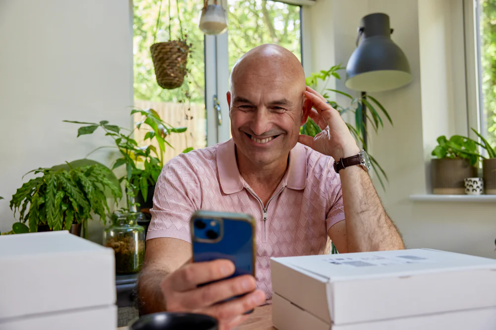 Customer sat at a desk holding their iPhone out in front of them and smiling in its direction with parcels stacked around them.