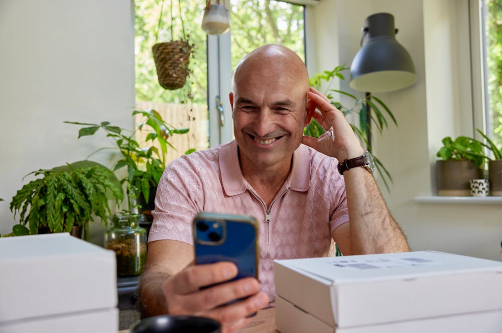 Customer sat at a desk holding their iPhone out in front of them and smiling in its direction with parcels stacked around them.