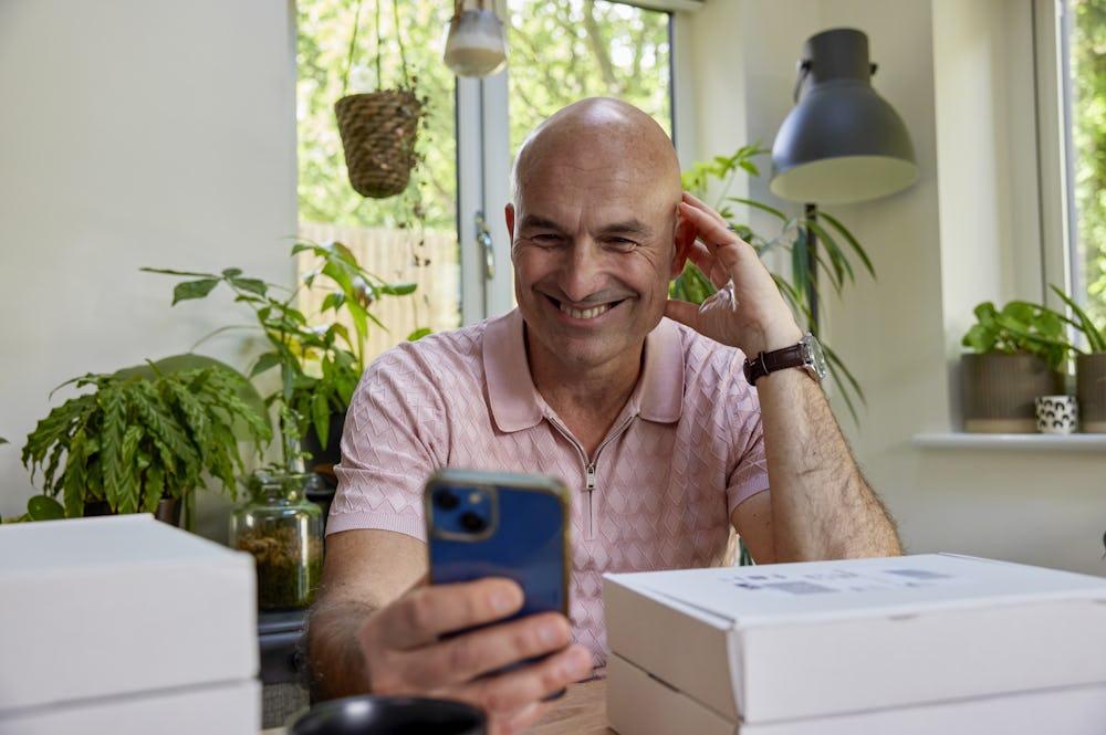 Customer sat at a desk holding their iPhone out in front of them and smiling in its direction with parcels stacked around them.