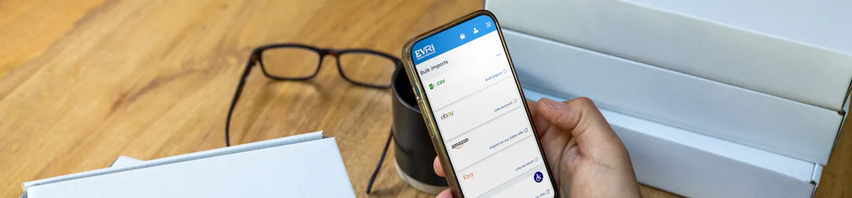 A mobile phone is held outright at a desk with parcels in the background. The screen displays Evri's website with the customer about to book their deliveries.
