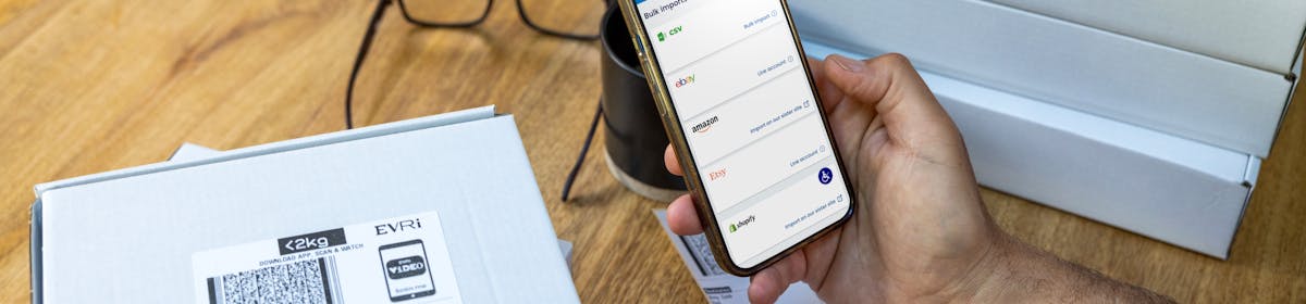 A mobile phone is held outright at a desk with parcels in the background. The screen displays Evri's website with the customer about to book their deliveries.