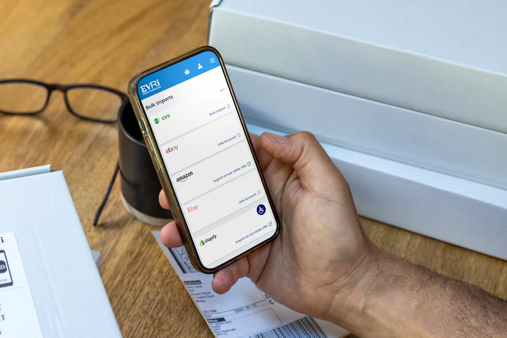 A mobile phone is held outright at a desk with parcels in the background. The screen displays Evri's website with the customer about to book their deliveries.