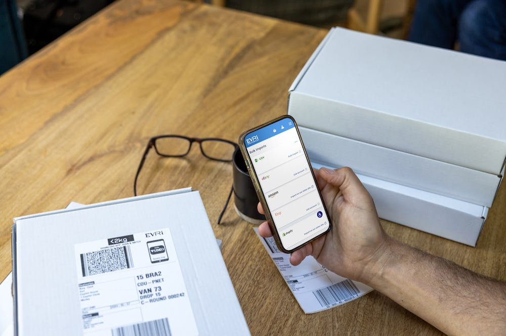 A mobile phone is held outright at a desk with parcels in the background. The screen displays Evri's website with the customer about to book their deliveries.