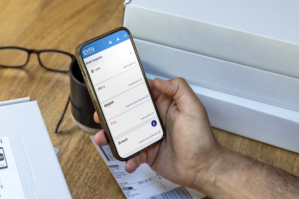 A mobile phone is held outright at a desk with parcels in the background. The screen displays Evri's website with the customer about to book their deliveries.