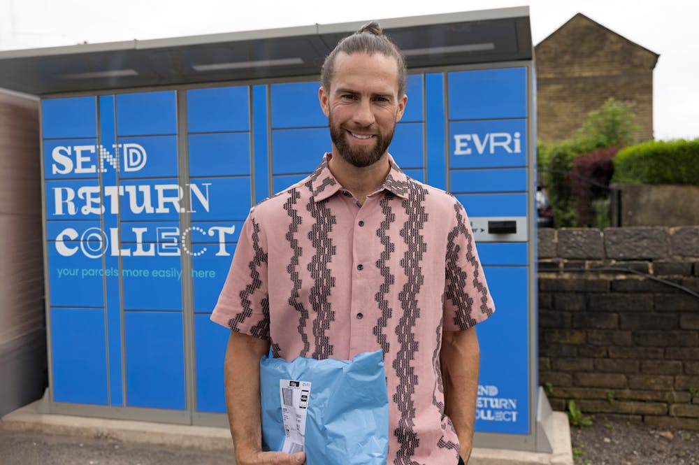 Customer standing outside with a blue Evri Locker in the background. They are holding a parcel and smiling for the photo.