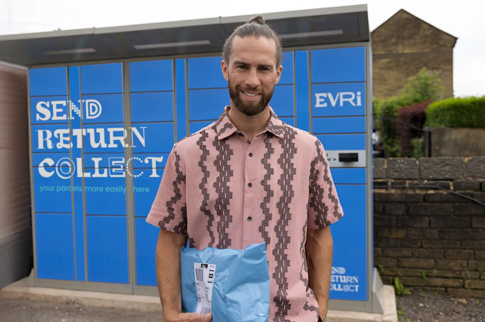 Customer standing outside with a blue Evri Locker in the background. They are holding a parcel and smiling for the photo.